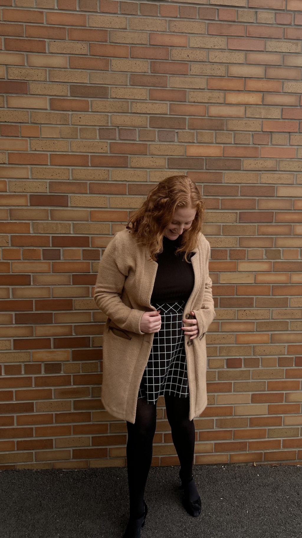 A woman smiles against brick wall while leaning forward to look down